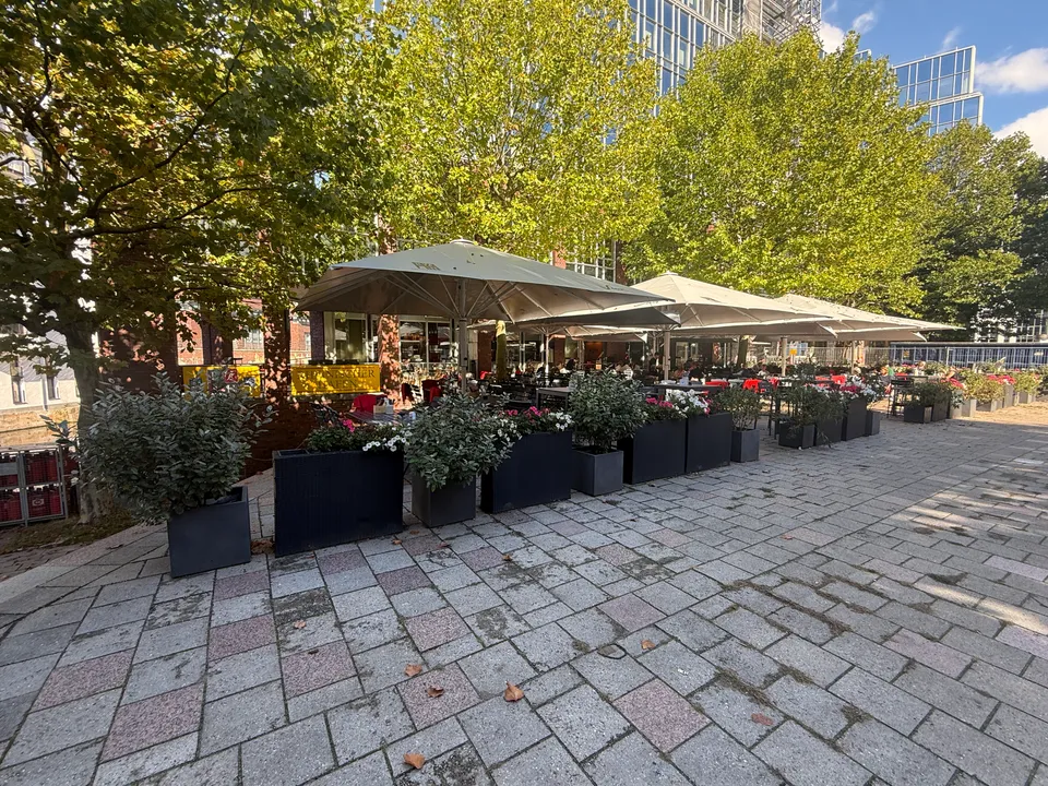 Restaurantterrasse mit beigen Sonnenschirmen und Blumenkübeln unter Platanen, davor ein gepflasterter Platz, dahinter Glasbürogebäude