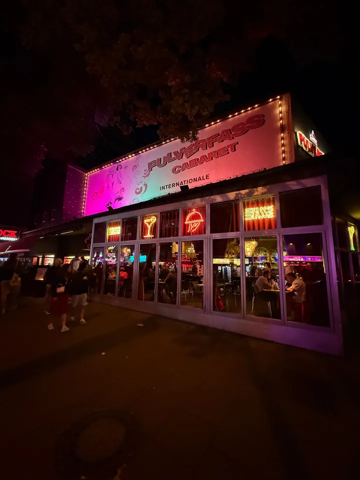 Pink beleuchtetes Transparent Pulverfass Cabaret über Glasfront mit Neonzeichen und Gästen, nachts auf dem Gehweg