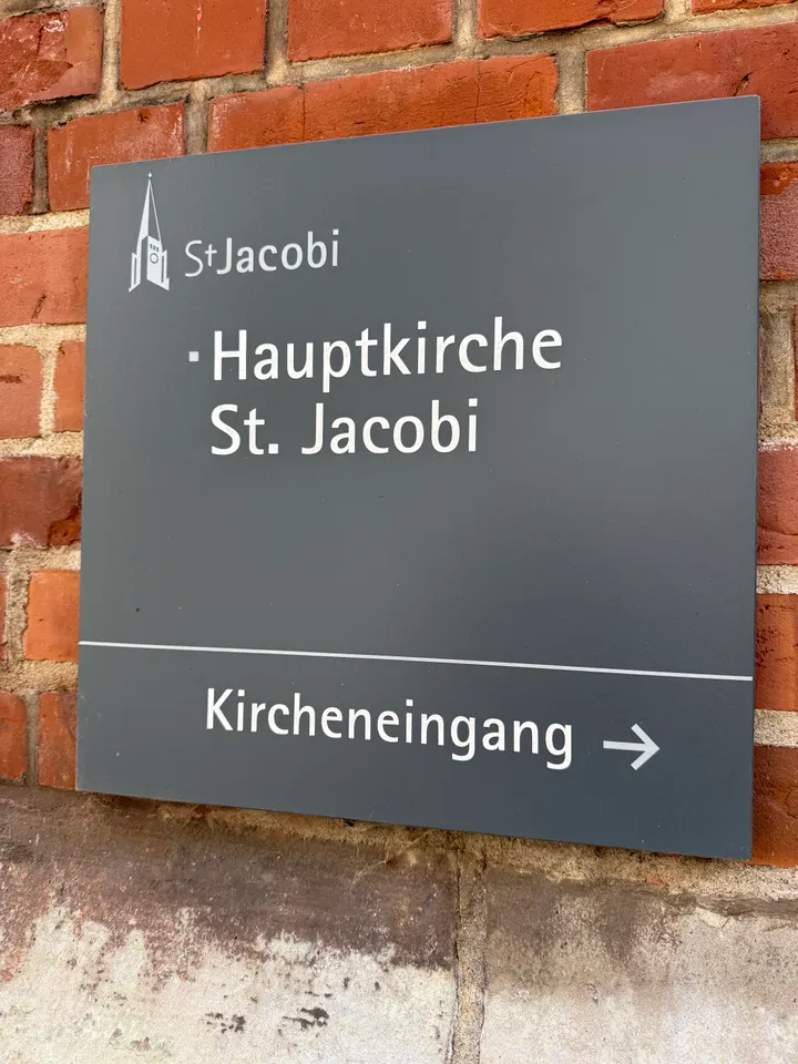 Graues Wandschild mit der Aufschrift Hauptkirche St. Jacobi und einem Pfeil zum Kircheneingang