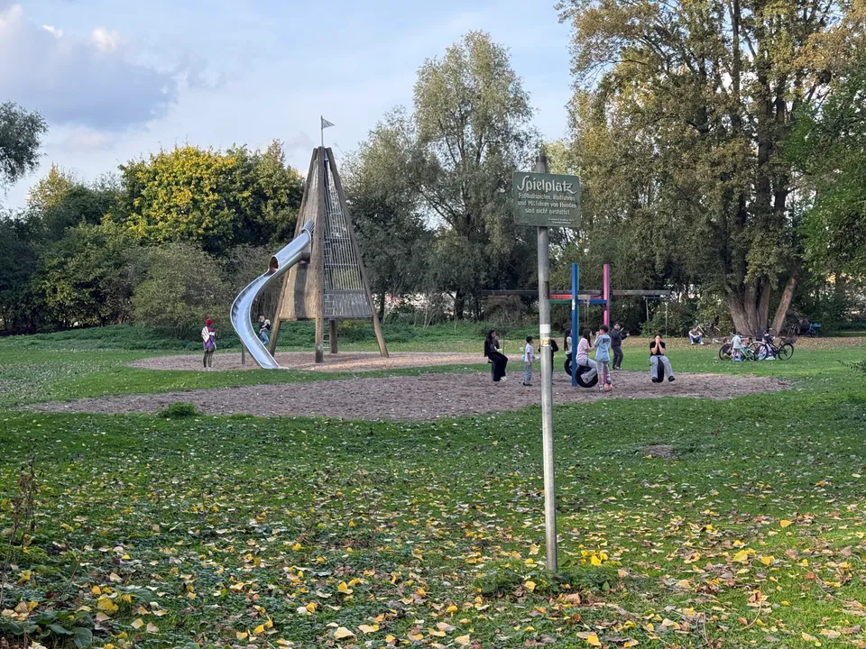 Spielplatz auf einer Parkwiese mit hölzernem Kletterturm, Edelstahlrutsche, bunten Reifenschaukeln und Spielplatzschild im Vordergrund