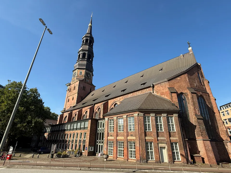 Backsteinkirche mit hohem Turm, gestufter Kupferhaube und zweigeschossigem Anbau an einer Straße unter blauem Himmel