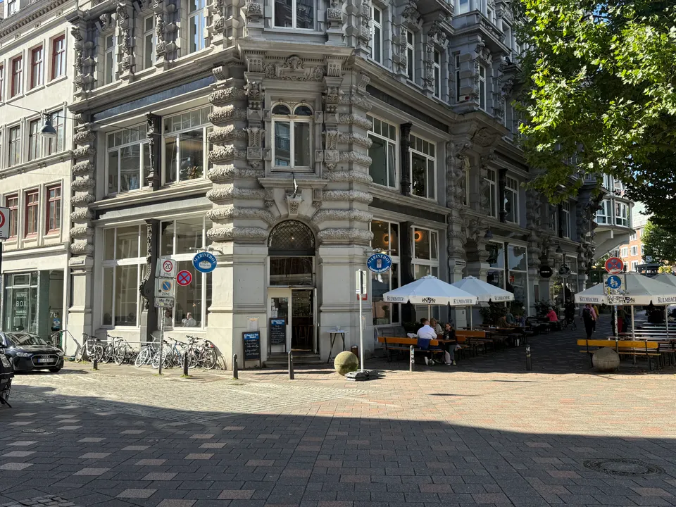 Verziertes Eckgebäude der Gaststätte Marinehof mit Sonnenschirmen, Biertischen und abgestellten Fahrrädern auf gepflastertem Platz