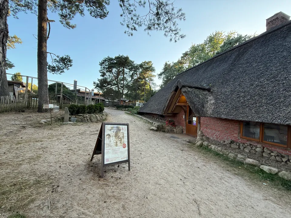 Sandweg neben reetgedecktem Backsteinhaus mit Holztür, davor Aufsteller mit Veranstaltungsplakat unter Kiefern