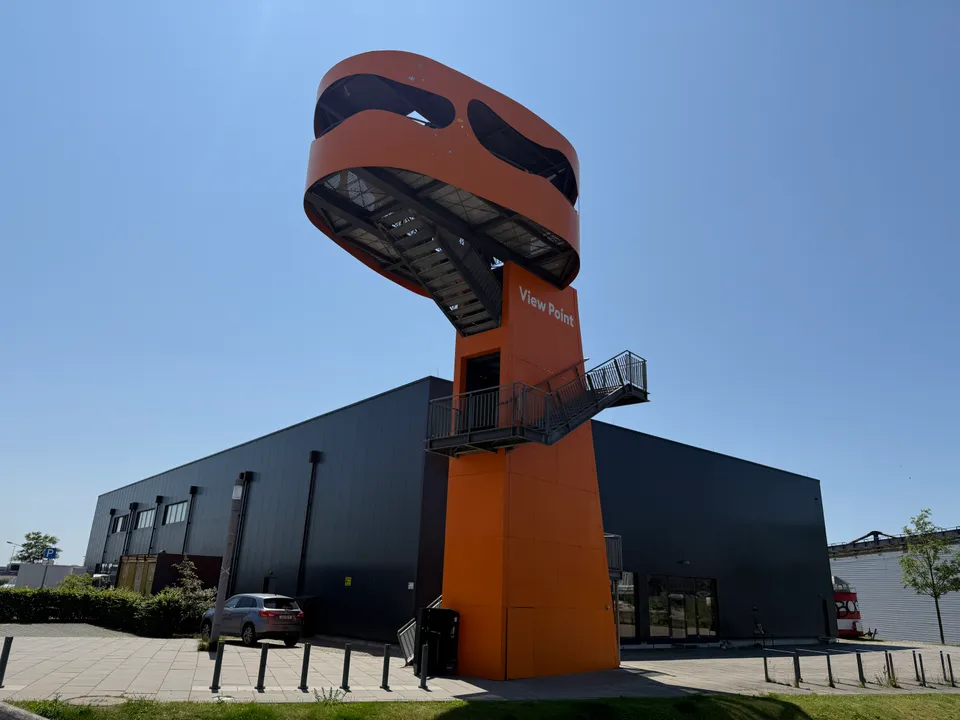 Orangefarbener Aussichtsturm mit ovaler Plattform und Schriftzug View Point vor dunkelgrauer Halle unter blauem Himmel