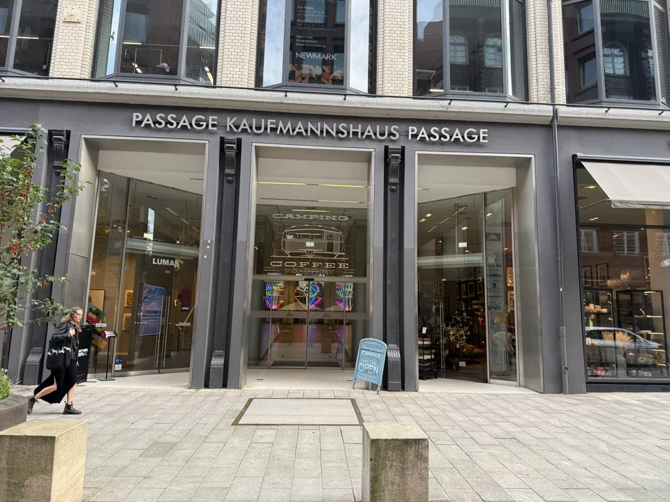 Eingang der Passage Kaufmannshaus mit dunkelgrauer Fassade, drei Glasportalen, Lumas-Schaufenster und Aufsteller eines Cafés