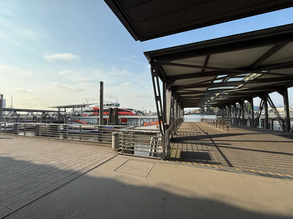 Überdachte Stahlbrücke zu einem Ponton an den Landungsbrücken, dahinter ein rot-weißer Katamaran an der Elbe