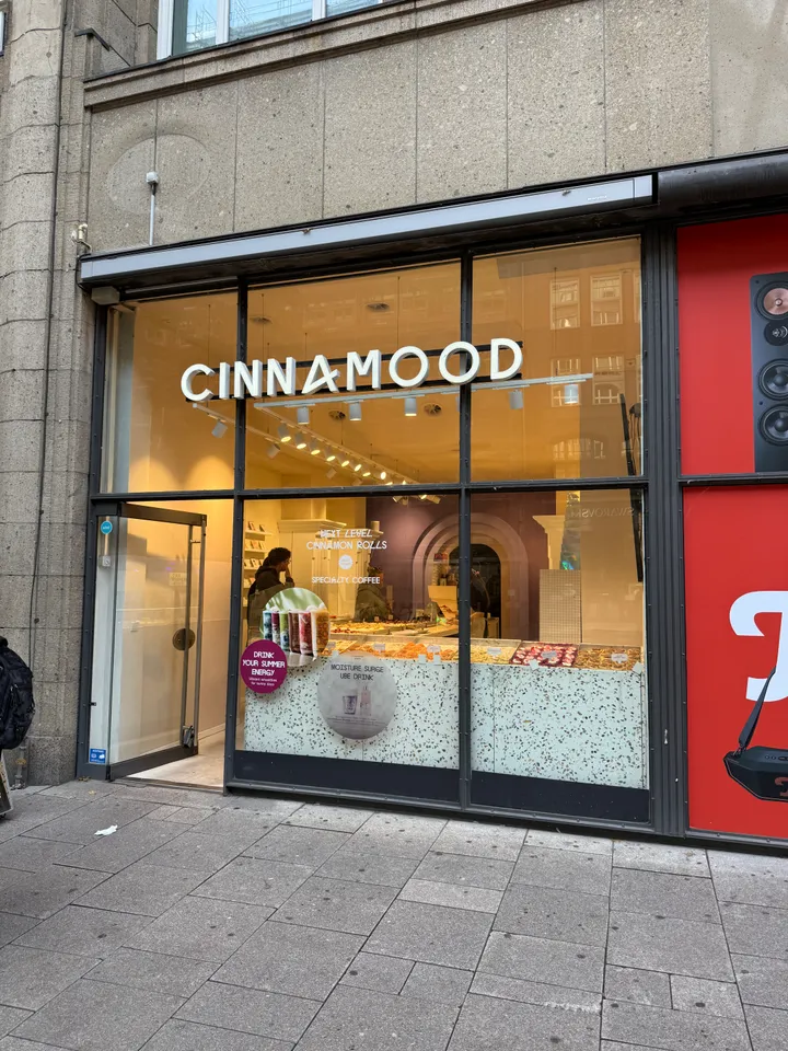Glasfront des Geschäfts Cinnamood mit weißem Schriftzug, geöffneter Tür und beleuchteter Gebäcktheke dahinter
