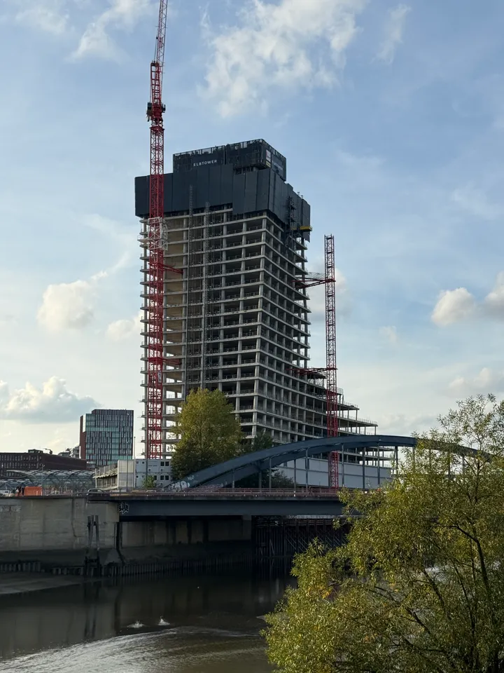 Hochhausrohbau mit Aufschrift Elbtower und roten Kränen hinter einer blauen Bogenbrücke über einem Gewässer