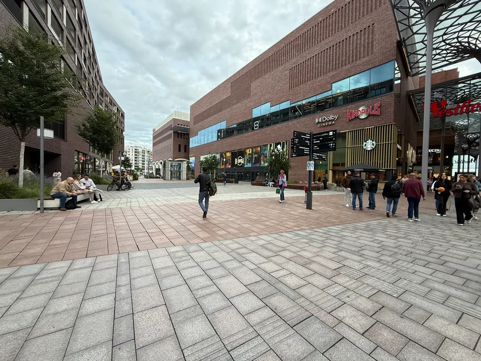 Gepflasterter Platz vor Klinkerbau mit Kinopolis-Kino, Starbucks, Wegweiser und Stahldach des Westfield-Einkaufszentrums, einige Fußgänger