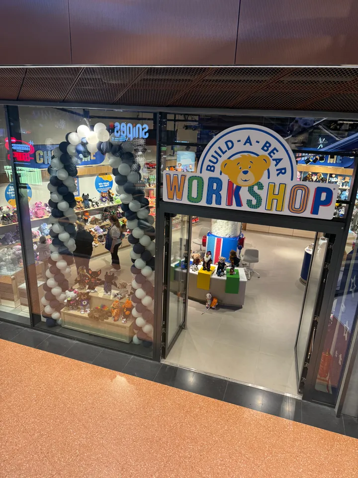 Eingang des Build-A-Bear-Workshop-Ladens schräg von oben, Ballonbogen im Schaufenster und Blick auf die Füllmaschine