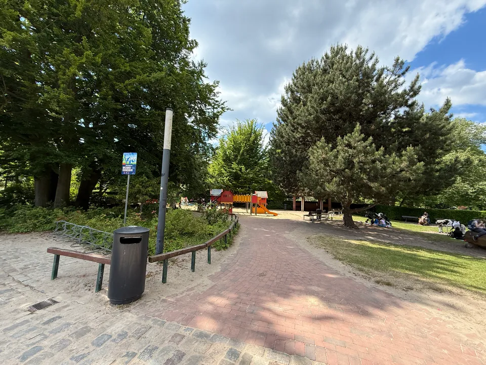 Klinkerweg zwischen großen Bäumen zu einem Spielhaus mit Rutsche, rechts Rasen mit Kinderwagen