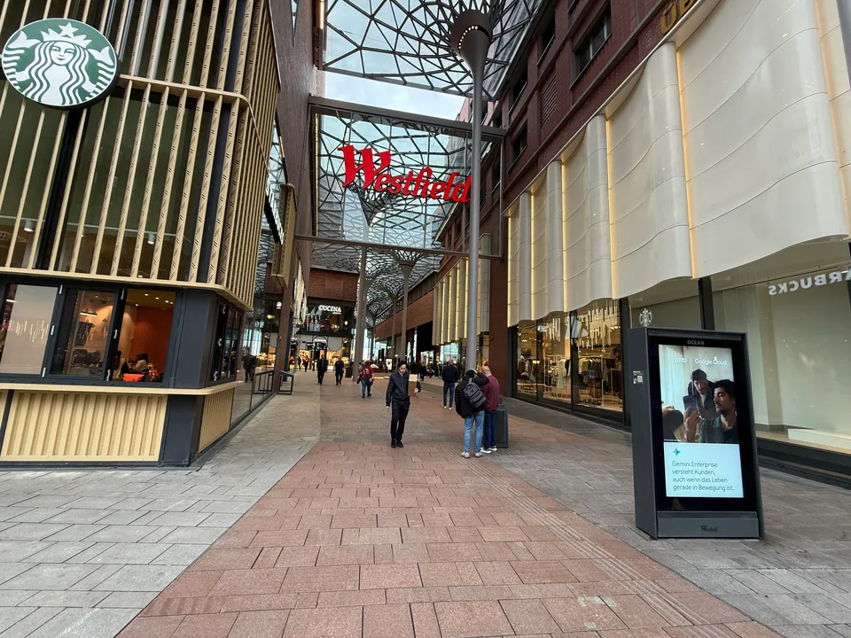 Überdachte Fußgängerpassage mit Glasdach und rotem Westfield-Schriftzug, links Starbucks-Filiale, rechts Backsteinfassade mit Geschäften