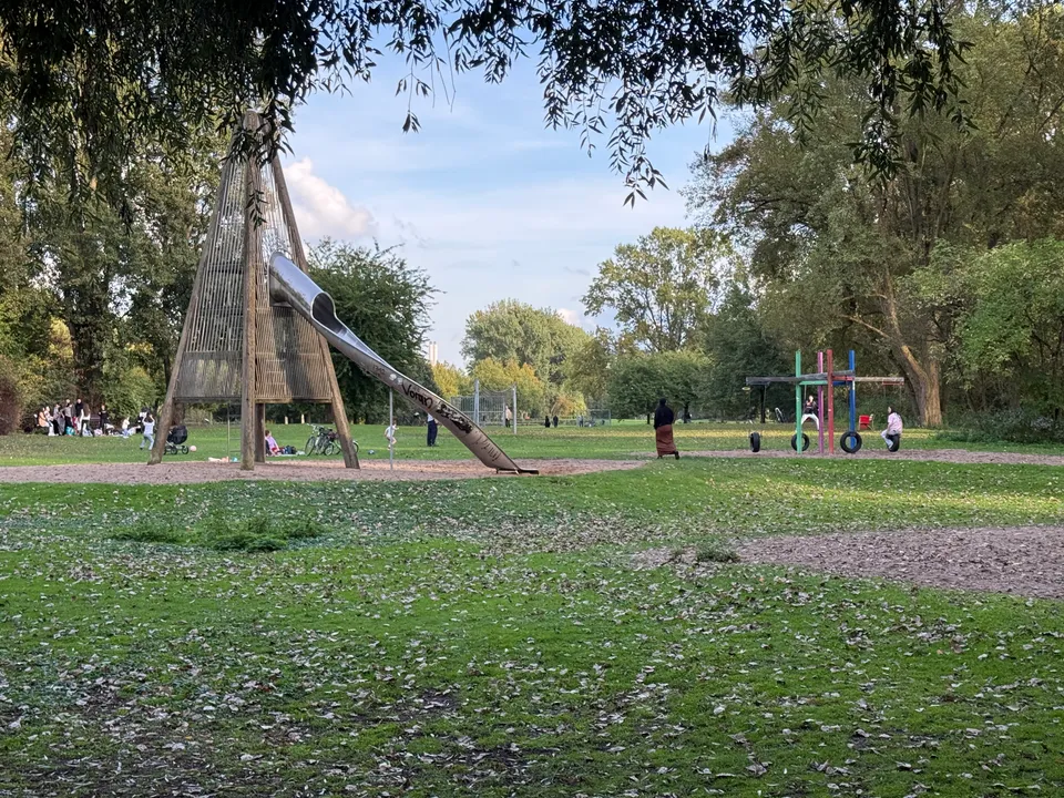 Parkwiese mit hölzernem Kletterturm und Edelstahlrutsche, rechts bunte Reifenschaukeln, eingerahmt von überhängenden Weidenzweigen