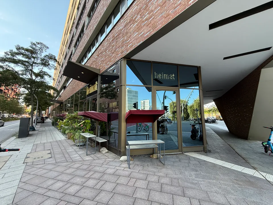 Verglaste Gebäudeecke mit Restaurant heimat, Außentischen unter roten Schirmdächern und Gehweg entlang einer Klinkerfassade bei blauem Himmel