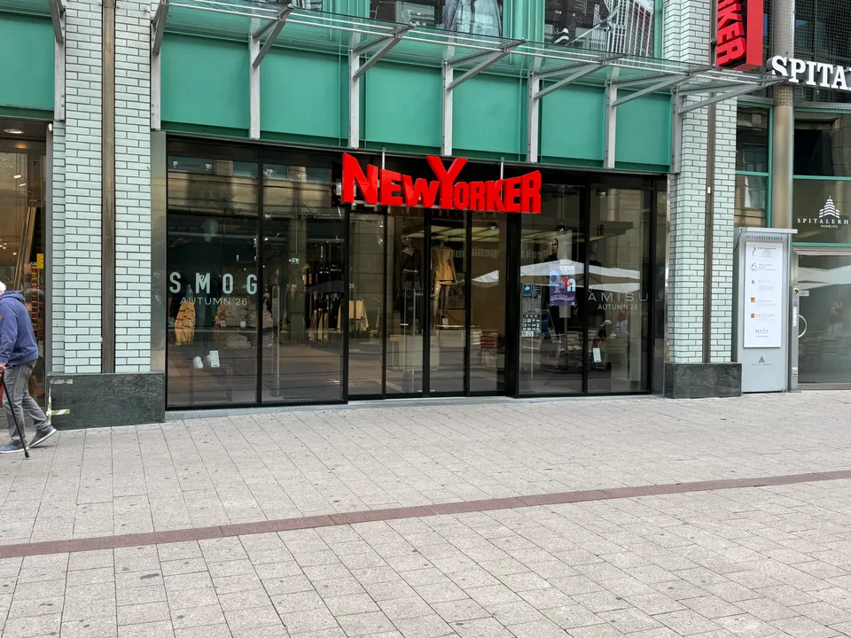 Ladenfront von New Yorker mit rotem Schriftzug, Glasschaufenstern und türkiser Fassade in einer Fußgängerzone