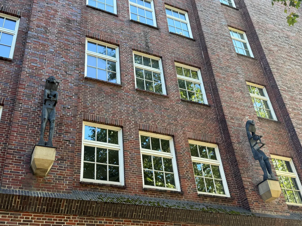 Zwei stilisierte Bronzefiguren auf Steinkonsolen an einer Klinkerfassade mit weißen Sprossenfenstern
