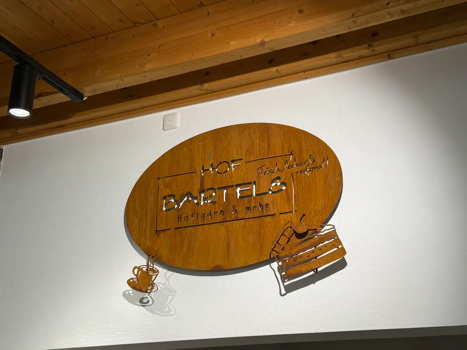 Rostbraunes ovales Metallschild mit der Aufschrift Hof Bartels, Hofladen & mehr, an einer weißen Wand