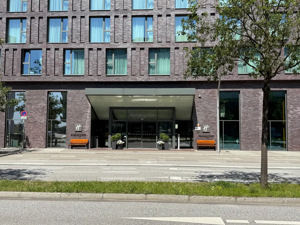 Hoteleingang mit gläsernem Vordach, Holiday-Inn-Logos, Blumenkübeln und orangefarbenen Bänken an dunkler Klinkerfassade