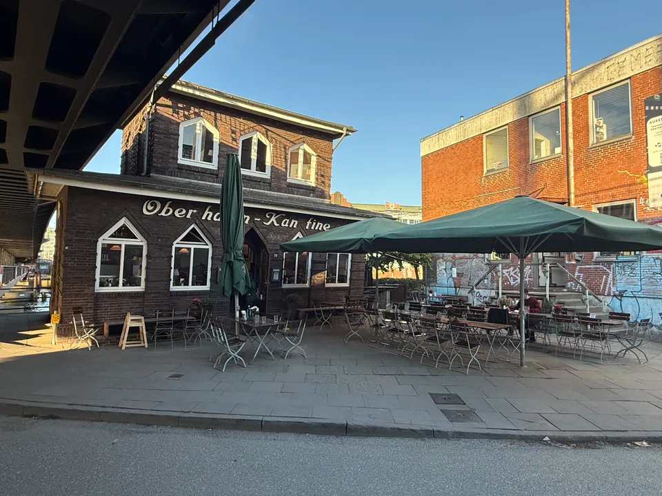 Oberhafen-Kantine mit Außenterrasse, Holztischen und grünen Sonnenschirmen unter einer Stahlbrücke