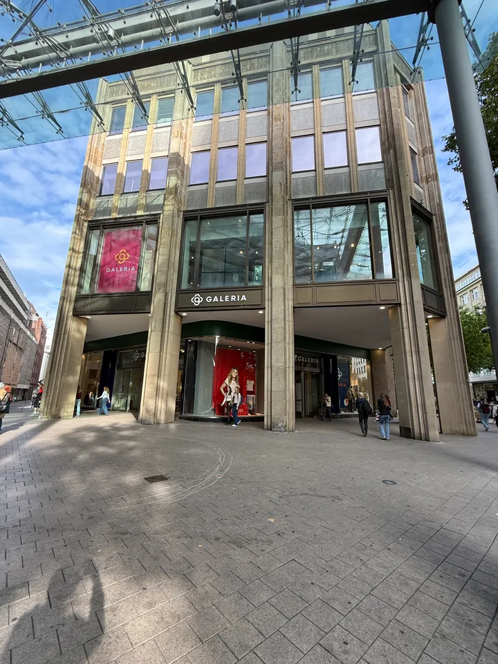 Kaufhausfassade mit pinkfarbenem GALERIA-Banner und Arkade, im Vordergrund gepflasterter Platz unter einem Glasdach