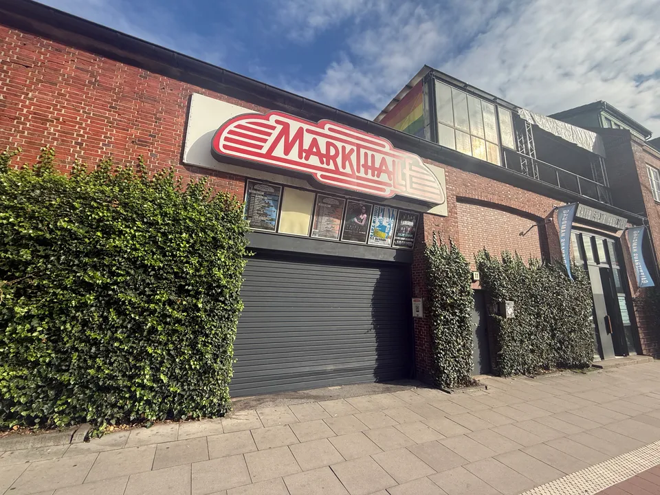 Backsteinfassade der Markthalle mit rot-weißem Schriftzug, Veranstaltungsplakaten, geschlossenem Rolltor und efeubewachsenen Wänden am Gehweg