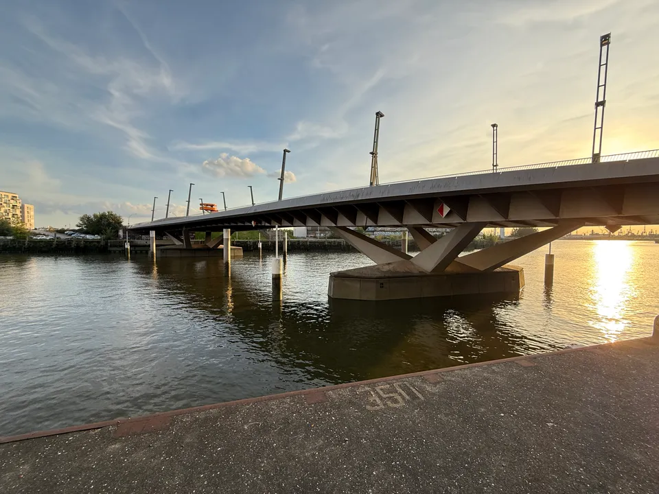 Flache Stahlbrücke mit V-förmigem Mittelpfeiler und hohen Lichtmasten über einem Hafenbecken bei tiefstehender Sonne