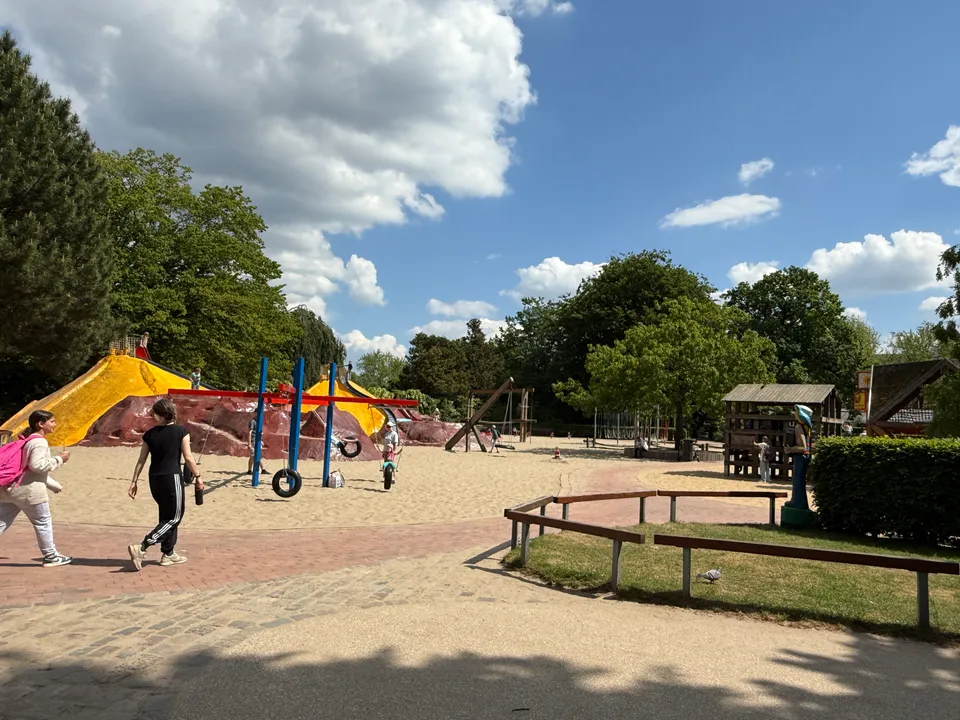 Sandspielplatz mit Reifenschaukel und gelben Kletterbergen, vorne zwei Fußgängerinnen auf dem Weg