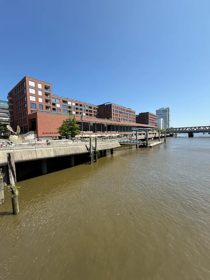Rote Klinkergebäude mit Arkaden an einer Hafenpromenade, davor Hafenbecken, rechts eine Stahlbrücke unter blauem Himmel