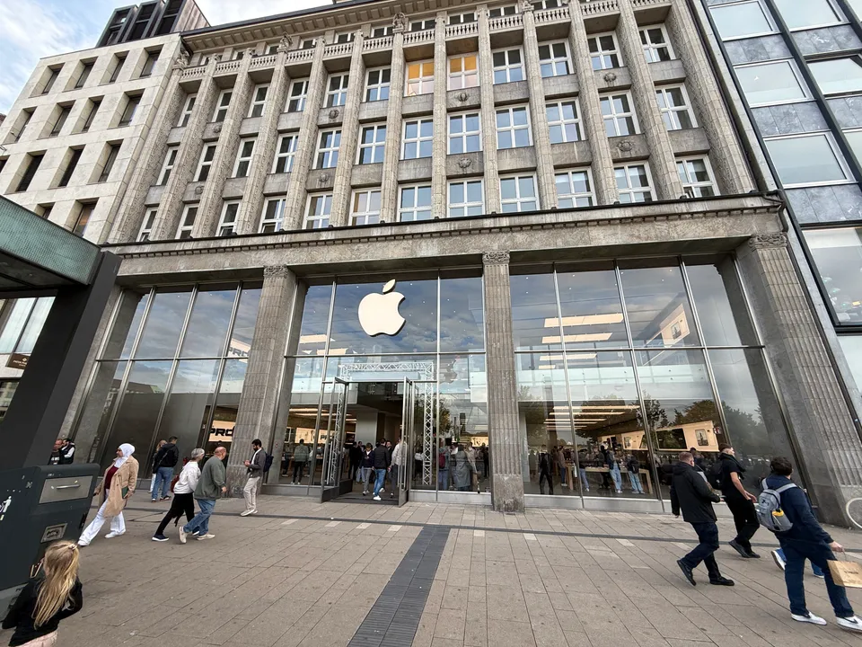 Steinfassade eines Geschäftshauses mit verglastem Erdgeschoss und Apple-Logo, davor Passanten auf breitem gepflastertem Gehweg