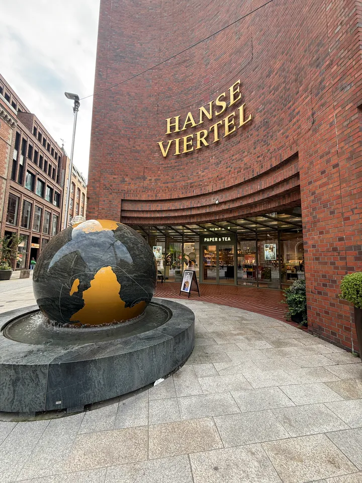 Globusbrunnen aus dunklem Stein vor geschwungenem Backsteineingang mit goldenem Schriftzug Hanse Viertel