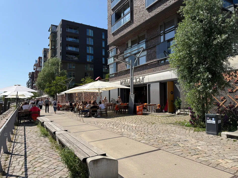 Gepflasterte Uferpromenade mit Holzbänken und voller Außenterrasse vor dem Lokal Kaisers in einem Klinkerbau