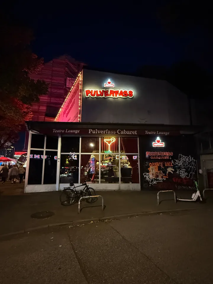 Nächtliche Fassade des Pulverfass Cabaret mit roter Leuchtschrift, Glasfront, Fahrrad und E-Scooter davor