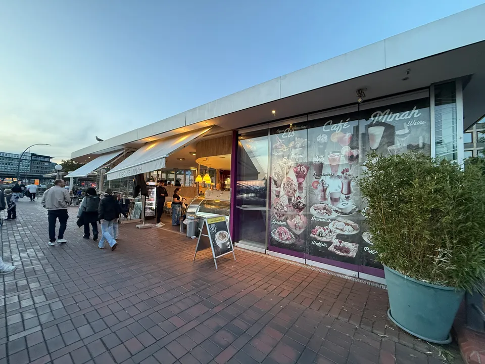 Eiscafé mit Markisen und beklebter Glasfront mit Eisbechern, davor Passanten auf Klinkerpflaster und ein Bambuskübel