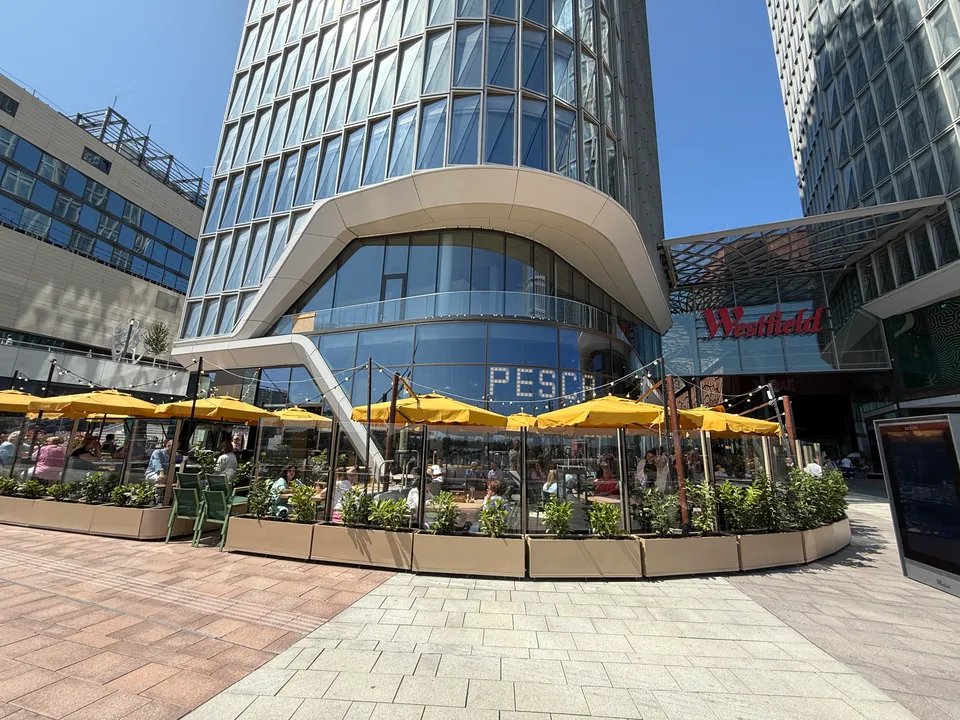 Restaurantterrasse mit gelben Sonnenschirmen vor einem verglasten Hochhaus, daneben der Eingang zum Westfield-Einkaufszentrum