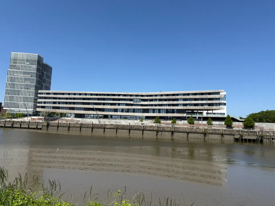 Langgestrecktes Gebäude mit hellen Fassadenbändern am Kai, daneben Glashochhaus, im Vordergrund Wasser und Ufergras