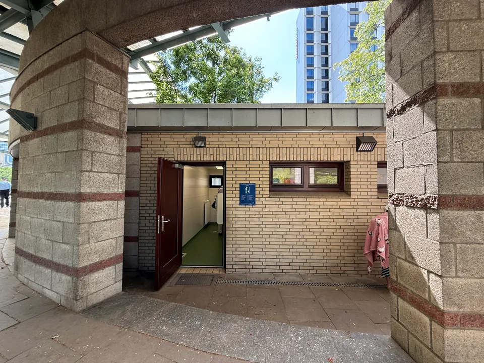 Toilettenhäuschen aus gelbem Klinker mit offener Tür zwischen gemauerten Pfeilern, dahinter ein Hochhaus