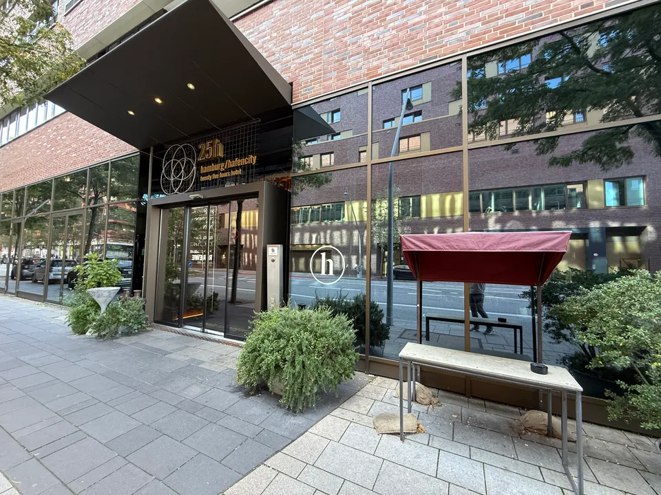 Eingang des 25hours Hotels HafenCity mit Schriftzug über der Glasschiebetür, Pflanzkübeln und Außentisch unter rotem Schirmdach