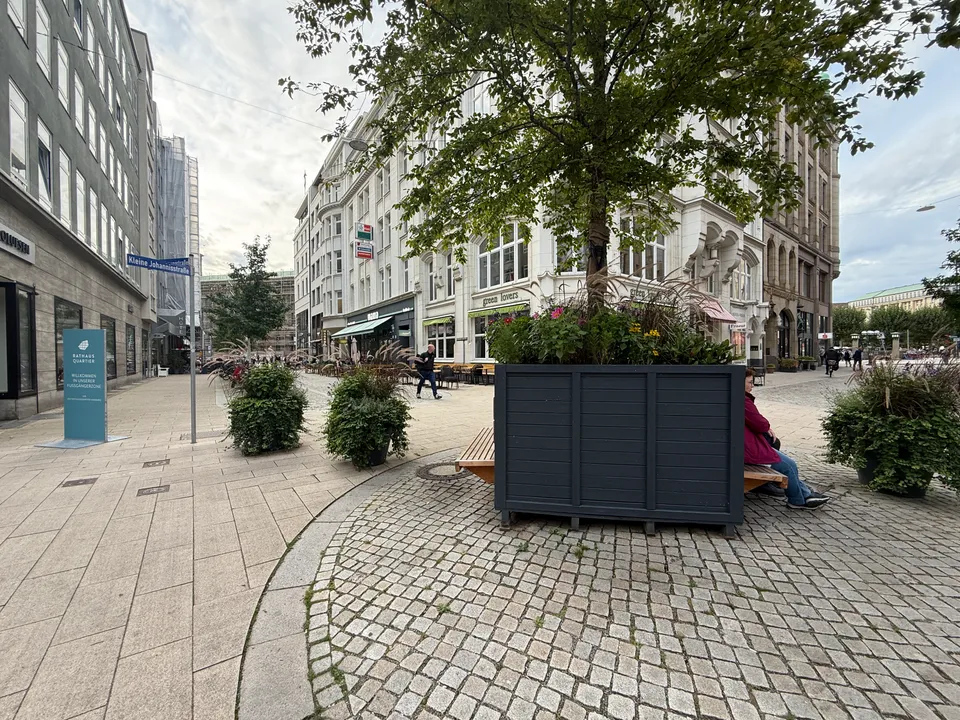 Holzpflanzkasten mit Baum und Sitzbänken auf Kopfsteinpflaster, dahinter helle Altbauten mit Cafés in der Fußgängerzone