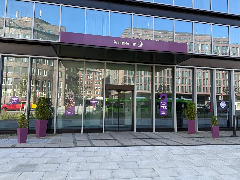 Verglaster Hoteleingang mit violettem Premier-Inn-Vordach, violetten Pflanzkübeln und gespiegelten Backsteingebäuden in der Fassade