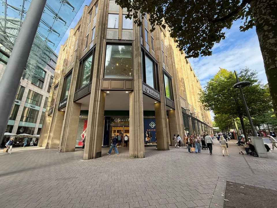 Natursteinfassade des GALERIA-Kaufhauses an einer Straßenecke, daneben Fußgängerzone mit Bäumen und Passanten