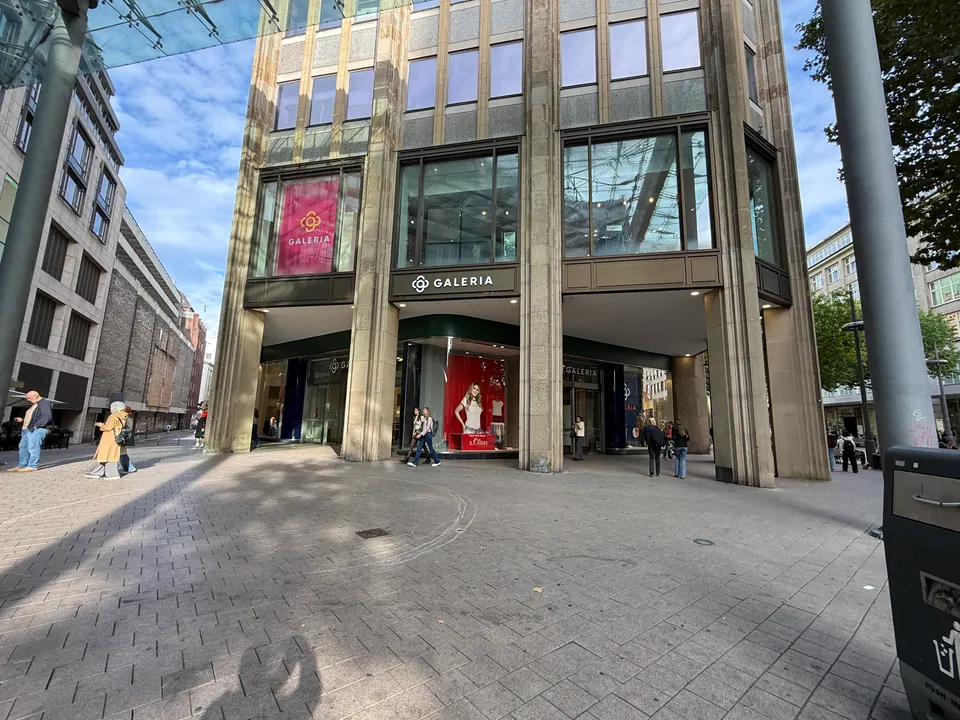 GALERIA-Kaufhaus mit Arkade, pinkfarbenem Banner und rotem Schaufenster, links Seitenstraße mit Backsteinbauten