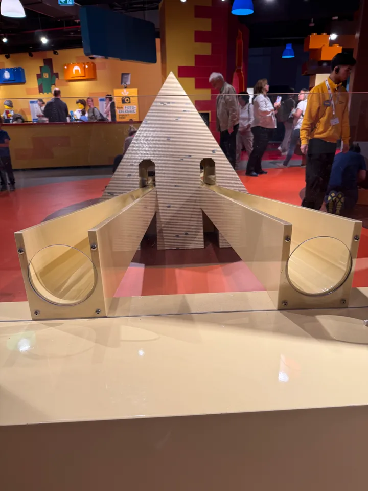 LEGO-Pyramide mit zwei Rollbahnen frontal in einer Vitrine, im Hintergrund Besucher und Spielhalle