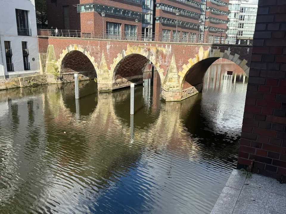 Historische Backsteinbrücke mit drei Sandsteinbögen über einem Fleet, spiegelt sich im Wasser, dahinter Backsteinbürogebäude