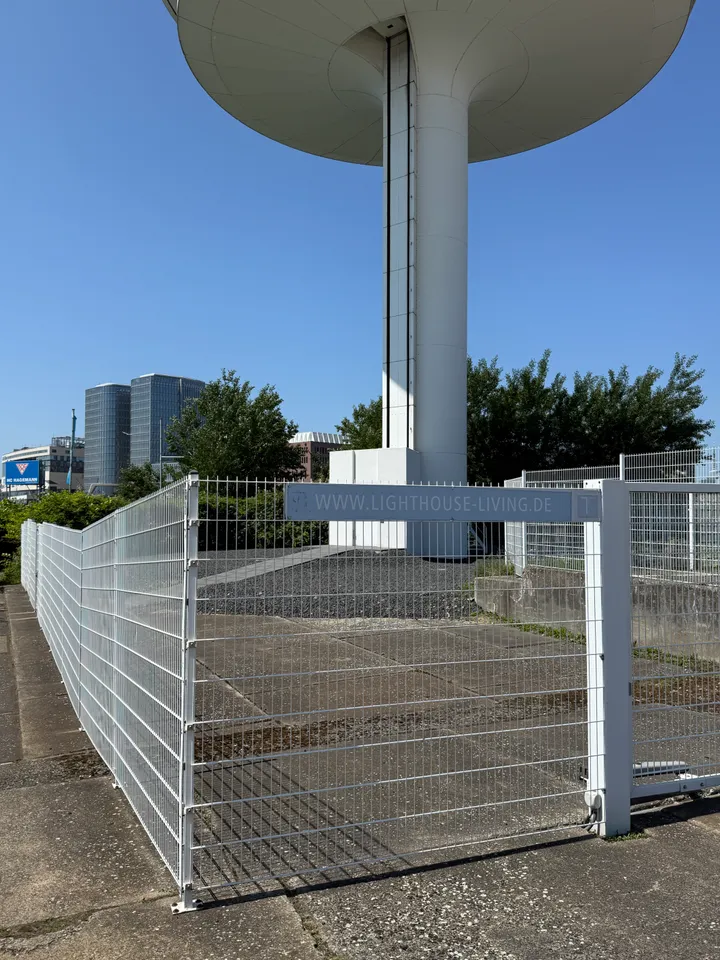 Weißer Gitterzaun mit Webadressenschild vor dem Sockel eines weißen Turms, links zwei verglaste Hochhäuser