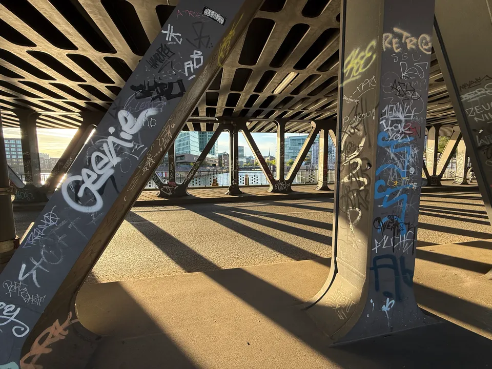 Graffitibedeckte Stahlstützen unter einer Brücke, dahinter Wasser und moderne Glasbürogebäude im Sonnenlicht