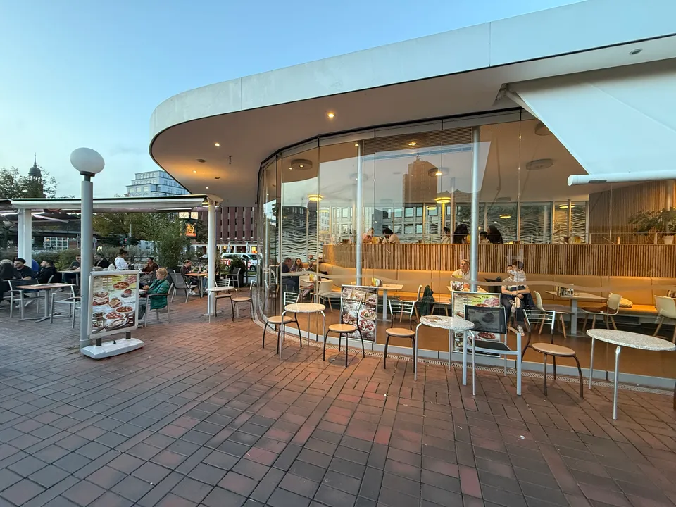 Pavillon mit geschwungener Glasfront und weißem Flachdach, davor Bistrotische auf Klinkerpflaster und Gäste auf der Außenterrasse