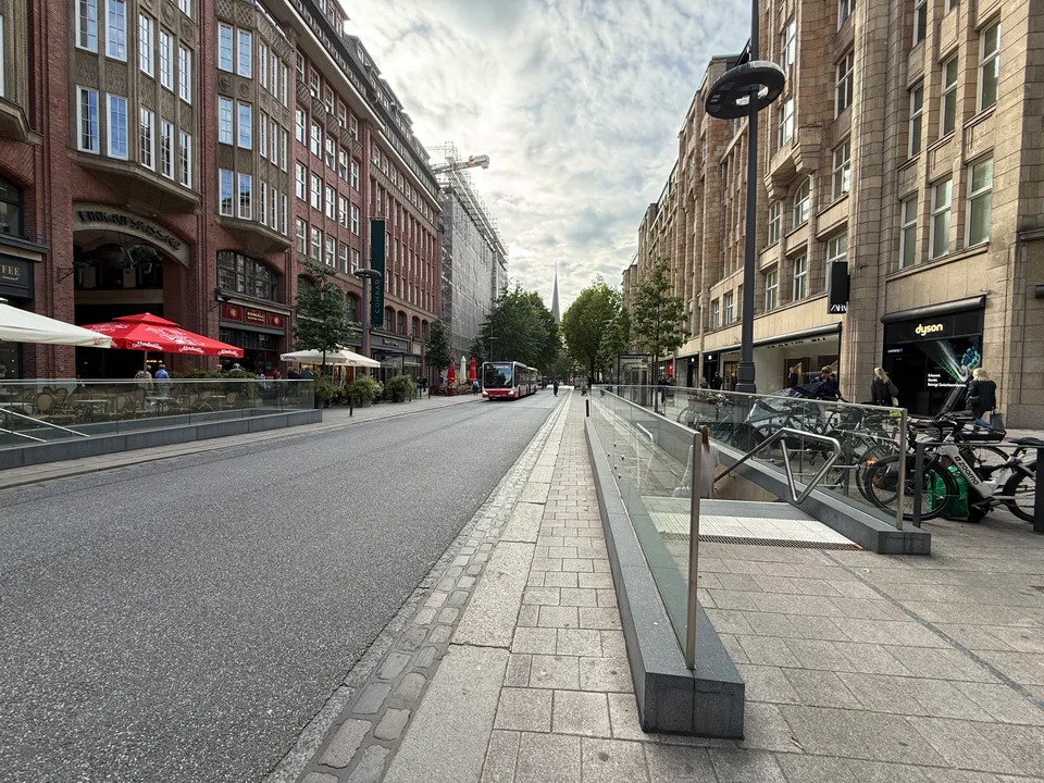 Straße mit Backstein- und Sandsteinfassaden, verglastem U-Bahn-Abgang, Straßencafé mit roten Schirmen und Kirchturm im Hintergrund