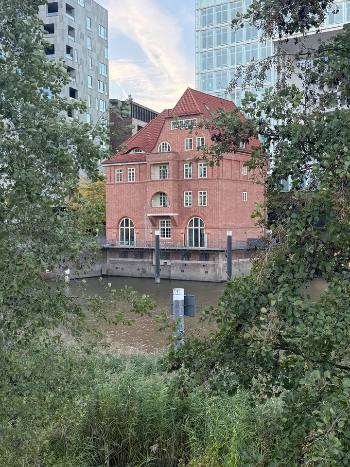 Rotes Backsteinhaus am Wasser, durch Baumäste und Schilf gesehen, im Hintergrund moderne Hochhäuser