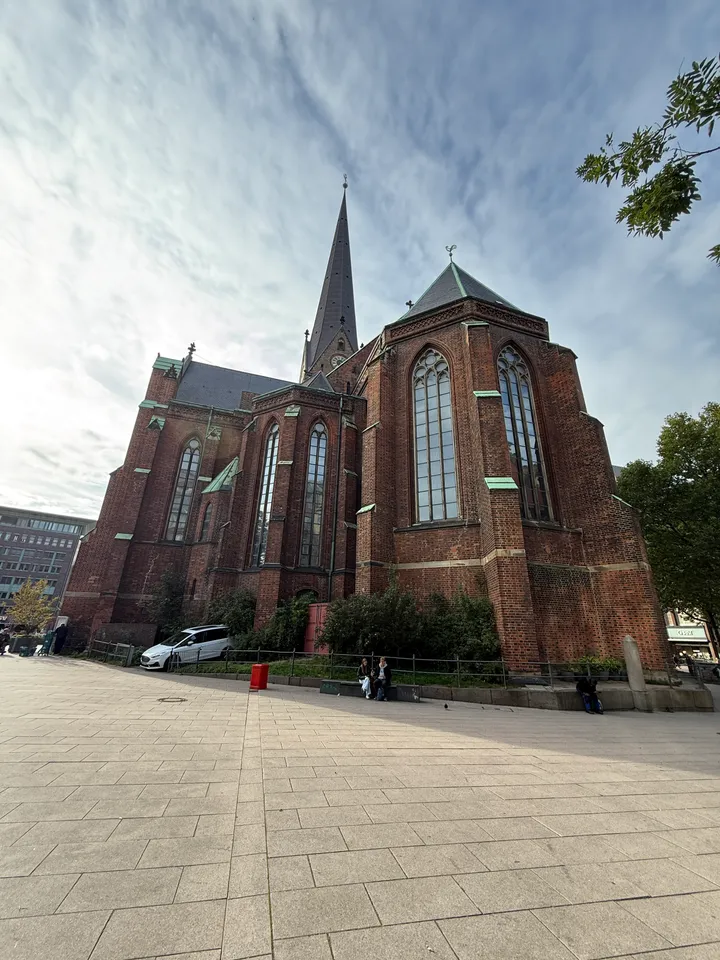 Chor einer roten Backsteinkirche mit hohen Spitzbogenfenstern und spitzem Turm hinter gepflastertem Platz