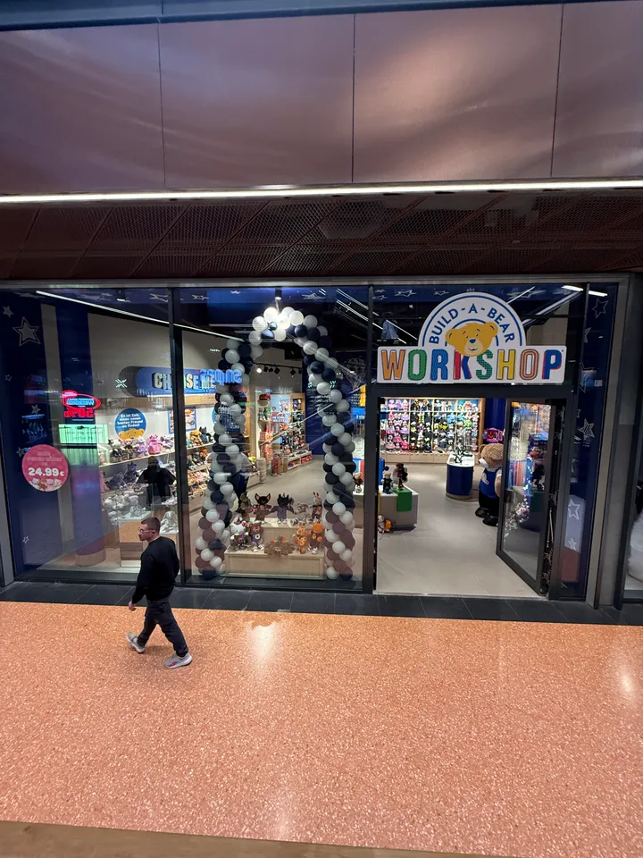 Glasfront des Build-A-Bear-Workshop-Ladens mit Ballonbogen im Schaufenster, davor ein Passant auf orangefarbenem Passagenboden